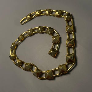 May include: A gold-toned necklace featuring a series of connected square links, each topped with a raised pyramid-shaped stud. The chain is arranged in a curved, open-ended shape and includes a hook-style clasp at one end for secure fastening.