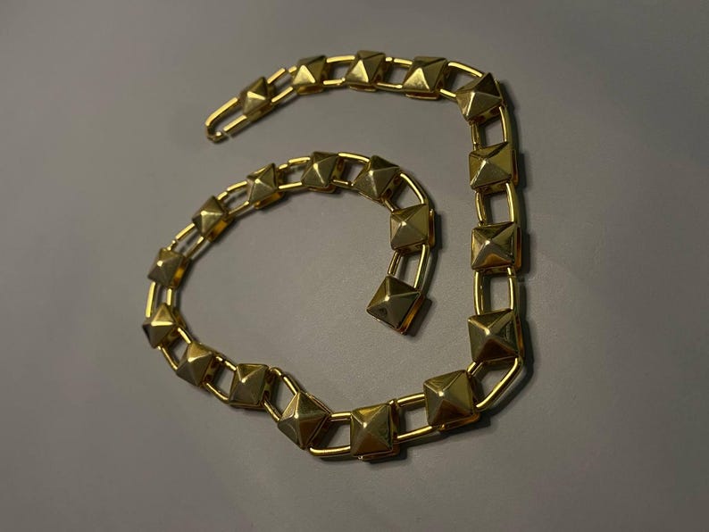 May include: A gold-toned necklace featuring a series of connected square links, each topped with a raised pyramid-shaped stud. The chain is arranged in a curved, open-ended shape and includes a hook-style clasp at one end for secure fastening.