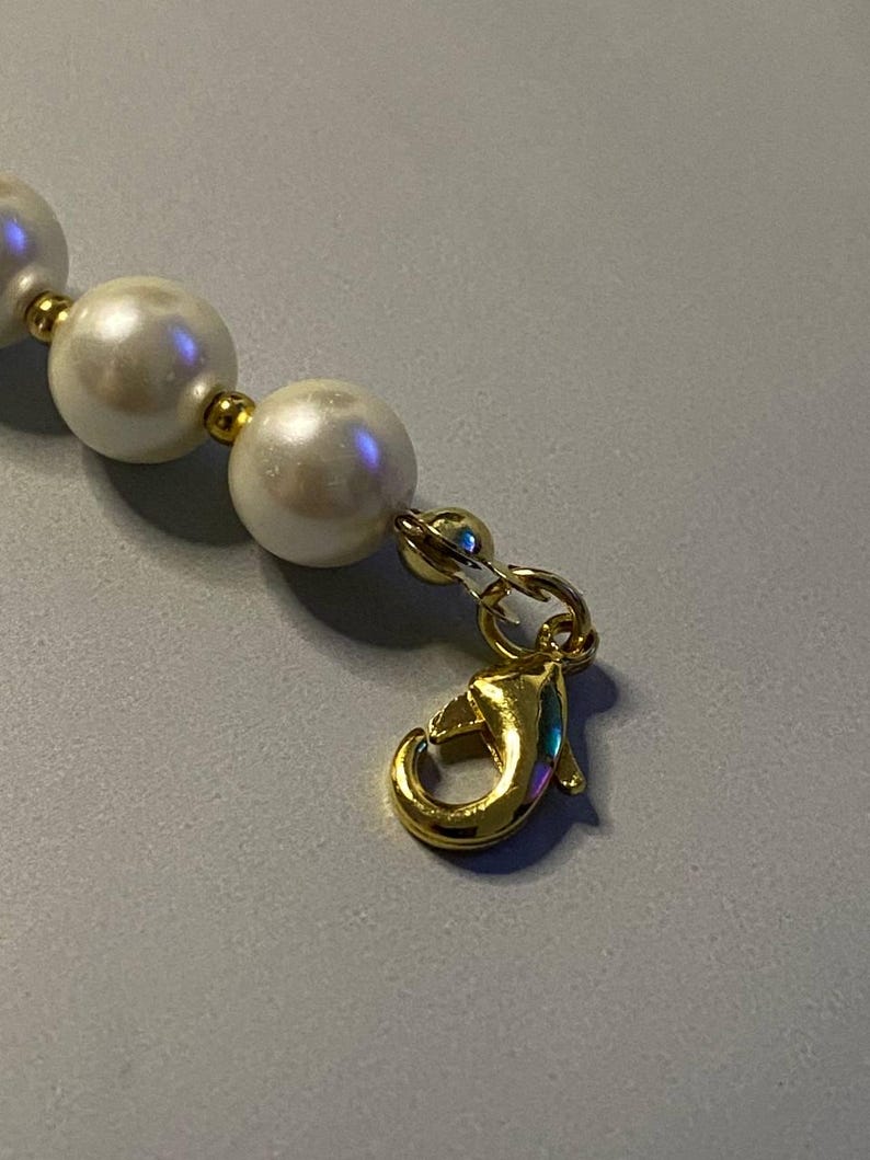 May include: A necklace featuring a series of round, white, lustrous beads separated by small, polished gold-tone metal spacers. The strand terminates with a gold-tone metal lobster claw clasp attached to a small jump ring, providing a secure closure.