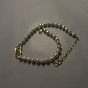 May include: A necklace featuring a strand of white spherical pearls interspersed with two gold-toned V-shaped metal charms. The piece includes a gold-toned metal chain extension and a lobster claw clasp, creating a circular design with metallic accents.
