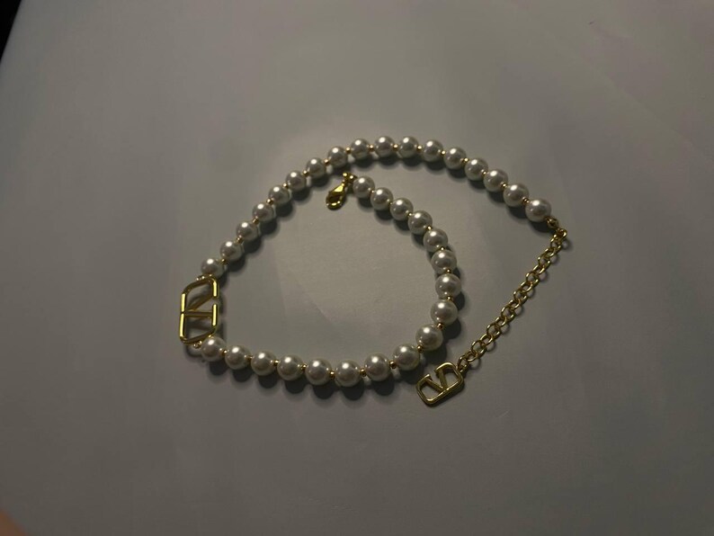 May include: A necklace featuring a strand of white spherical pearls interspersed with two gold-toned V-shaped metal charms. The piece includes a gold-toned metal chain extension and a lobster claw clasp, creating a circular design with metallic accents.