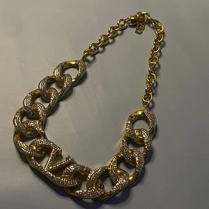 Puede incluir: Un collar de cadena dorada con eslabones ovalados grandes, gruesos y entrelazados. Varios de los eslabones est&aacute;n incrustados con peque&ntilde;os cristales claros y brillantes en sus caras frontales, mientras que los dem&aacute;s son de metal dorado liso y pulido.