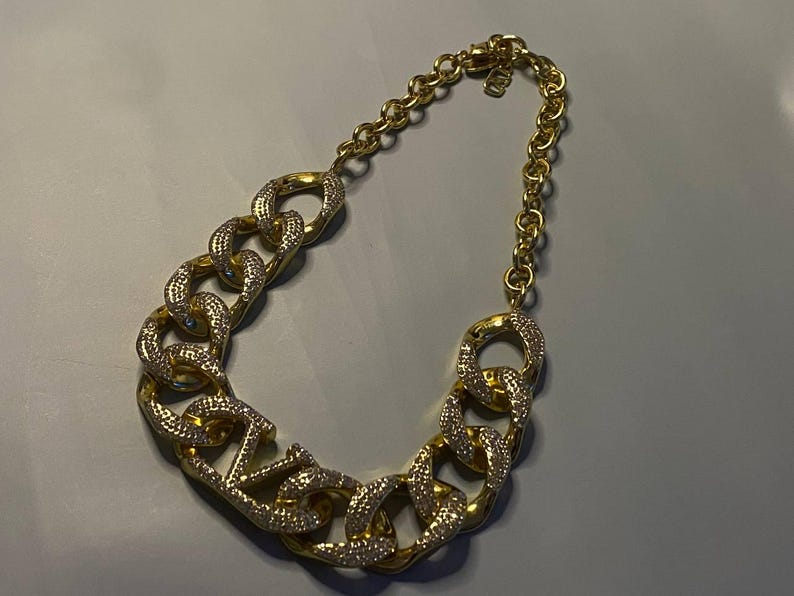 Puede incluir: Un collar de cadena dorada con eslabones ovalados grandes, gruesos y entrelazados. Varios de los eslabones est&aacute;n incrustados con peque&ntilde;os cristales claros y brillantes en sus caras frontales, mientras que los dem&aacute;s son de metal dorado liso y pulido.