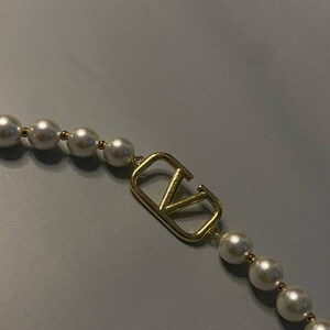 May include: A necklace featuring a series of round white pearls separated by small gold-toned beads, centered with a gold-toned metal V-shaped logo emblem. The design incorporates a repeating pattern of pearls and metallic accents along the strand.