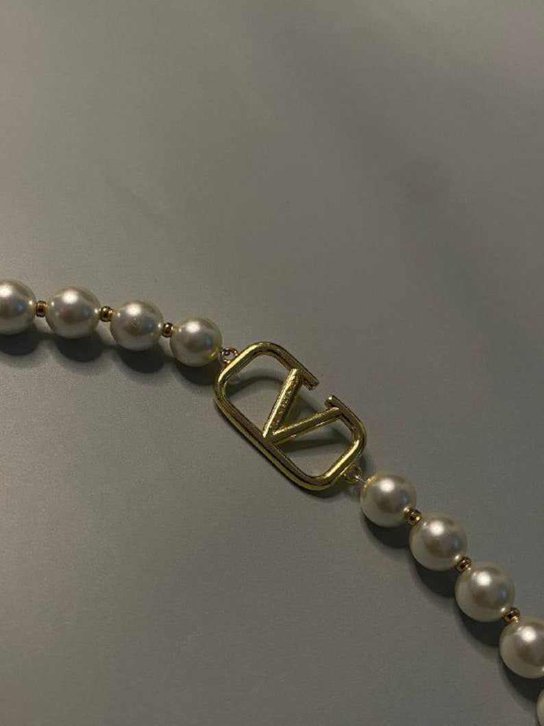 May include: A necklace featuring a series of round white pearls separated by small gold-toned beads, centered with a gold-toned metal V-shaped logo emblem. The design incorporates a repeating pattern of pearls and metallic accents along the strand.