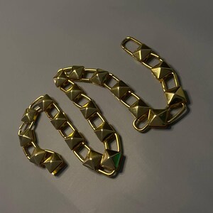 May include: A gold-toned chain strap featuring a series of connected square links, each topped with a raised pyramid-shaped stud. The chain is flexible and includes a rectangular clasp at one end, designed for attachment to a bag or accessory.