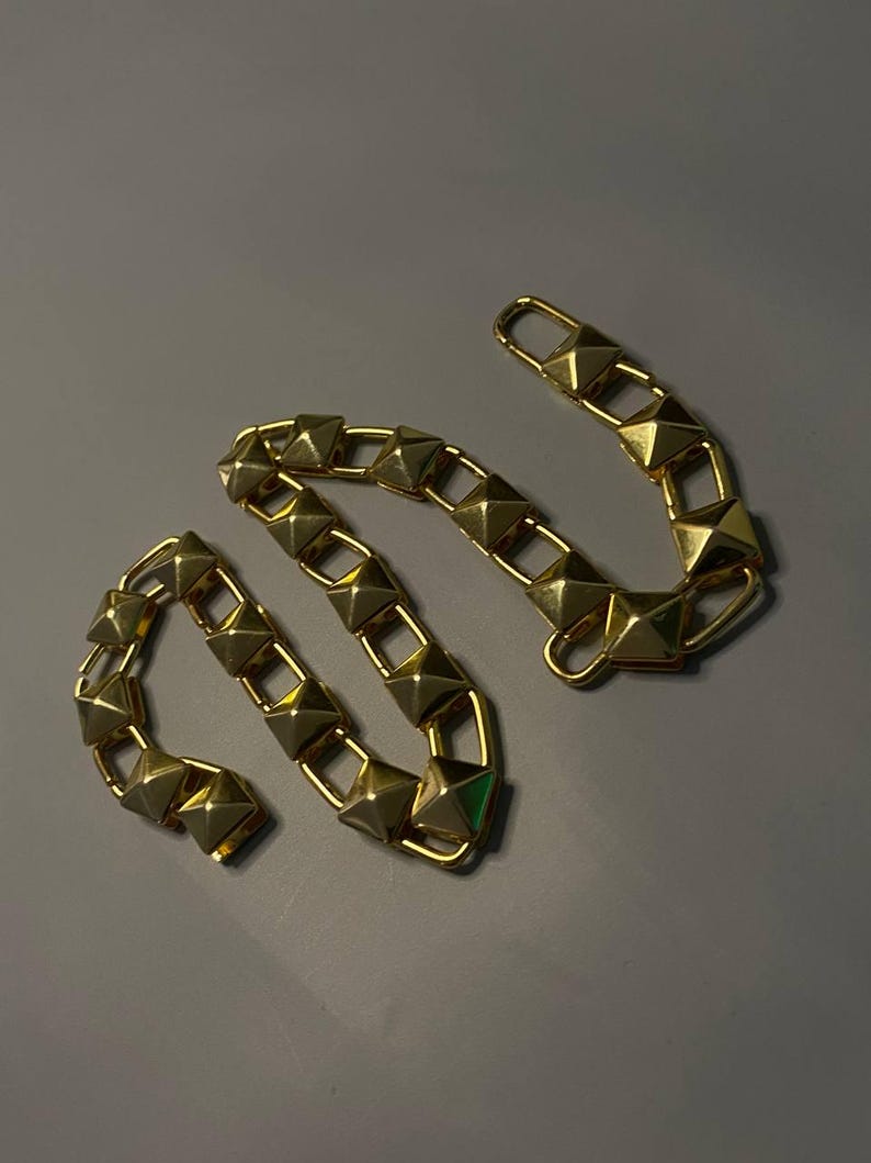 May include: A gold-toned chain strap featuring a series of connected square links, each topped with a raised pyramid-shaped stud. The chain is flexible and includes a rectangular clasp at one end, designed for attachment to a bag or accessory.