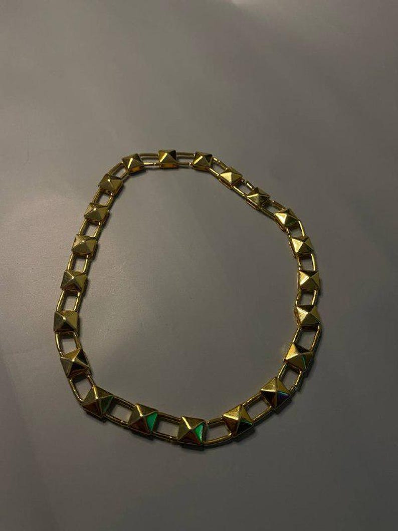 May include: A gold-toned chain necklace featuring a series of repeating pyramid-shaped studs linked together. The metallic links form a circular shape, with each pyramid stud displaying a smooth, reflective surface and a pointed top.