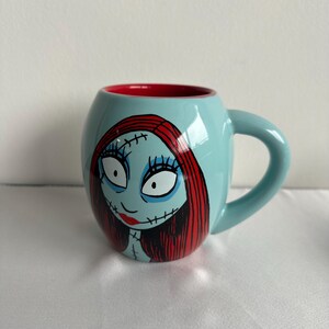 Pode incluir: Uma caneca de cer&acirc;mica azul-clara com formato arredondado e interior vermelho. O exterior apresenta uma ilustra&ccedil;&atilde;o gr&aacute;fica de uma personagem com longos cabelos vermelhos, olhos brancos grandes e detalhes de costura preta no rosto e pesco&ccedil;o.