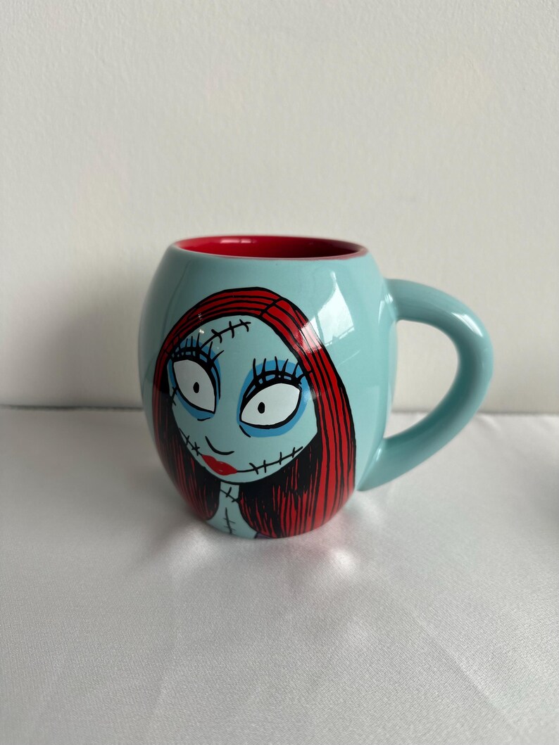 Pode incluir: Uma caneca de cer&acirc;mica azul-clara com formato arredondado e interior vermelho. O exterior apresenta uma ilustra&ccedil;&atilde;o gr&aacute;fica de uma personagem com longos cabelos vermelhos, olhos brancos grandes e detalhes de costura preta no rosto e pesco&ccedil;o.