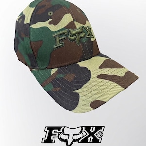 May include: A camouflage baseball cap featuring a green, brown, and tan pattern. The front panel displays the embroidered text FOX with a stylized fox head logo in the center. The cap has a curved brim and stitched eyelets on the crown.