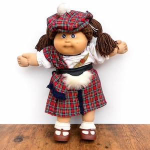 May include: A Cabbage Patch Kids doll with brown yarn hair in pigtails, wearing a red and green plaid tam hat with a white pom-pom, a white blouse, a black belt with a white faux fur sporran, a matching plaid kilt, and brown shoes with white socks.