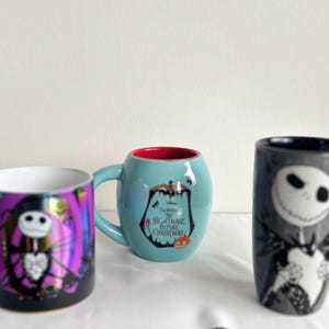 Pode incluir: Tr&ecirc;s canecas de cer&acirc;mica com personagens de O Estranho Mundo de Jack. Uma caneca &eacute; roxa com um gr&aacute;fico do Jack Skellington, uma &eacute; azul-clara com interior vermelho e logo do filme, e uma &eacute; alta, cinza e tem uma al&ccedil;a listrada.