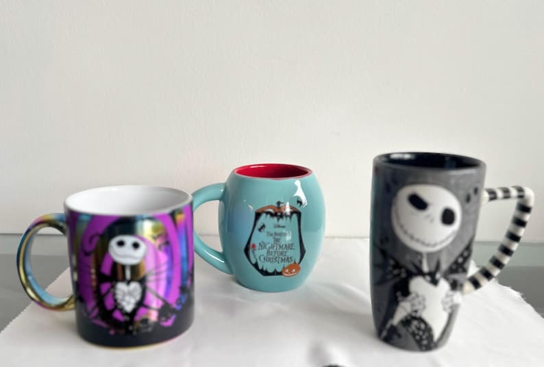 Pode incluir: Tr&ecirc;s canecas de cer&acirc;mica com personagens de O Estranho Mundo de Jack. Uma caneca &eacute; roxa com um gr&aacute;fico do Jack Skellington, uma &eacute; azul-clara com interior vermelho e logo do filme, e uma &eacute; alta, cinza e tem uma al&ccedil;a listrada.