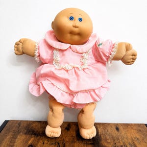 Pode incluir: Uma boneca careca com cabeça redonda, olhos azuis e corpo de tecido macio. A boneca veste um vestido rosa claro com gola arredondada, mangas curtas bufantes com acabamento em renda e bainha com babados. Uma pequena etiqueta verde é visível na manga esquerda.