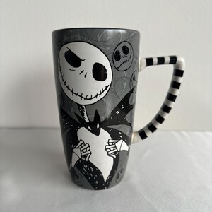 Pode incluir: Uma caneca alta de cer&acirc;mica com fundo cinza e uma grande ilustra&ccedil;&atilde;o em preto e branco de um personagem esquel&eacute;tico com a boca costurada. A caneca tem uma al&ccedil;a listrada em preto e branco e rostos circulares menores no fundo.