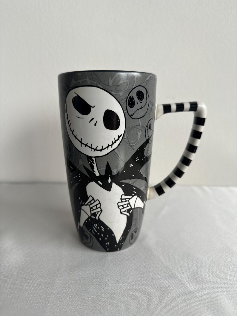Pode incluir: Uma caneca alta de cer&acirc;mica com fundo cinza e uma grande ilustra&ccedil;&atilde;o em preto e branco de um personagem esquel&eacute;tico com a boca costurada. A caneca tem uma al&ccedil;a listrada em preto e branco e rostos circulares menores no fundo.
