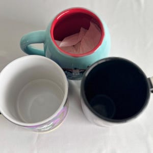 Pode incluir: Tr&ecirc;s canecas de cer&acirc;mica vistas de cima. Uma caneca &eacute; azul-clara com interior vermelho, uma &eacute; branca com uma al&ccedil;a colorida de arco-&iacute;ris e uma &eacute; preta com uma al&ccedil;a listrada de preto e branco. As canecas est&atilde;o dispostas em forma&ccedil;&atilde;o triangular.