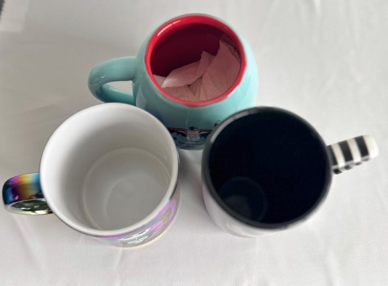 Pode incluir: Tr&ecirc;s canecas de cer&acirc;mica vistas de cima. Uma caneca &eacute; azul-clara com interior vermelho, uma &eacute; branca com uma al&ccedil;a colorida de arco-&iacute;ris e uma &eacute; preta com uma al&ccedil;a listrada de preto e branco. As canecas est&atilde;o dispostas em forma&ccedil;&atilde;o triangular.