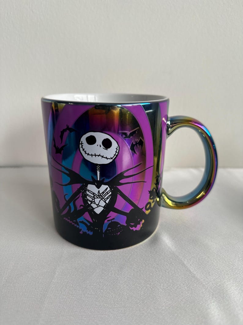 Pode incluir: Uma caneca de cer&acirc;mica com acabamento iridescente multicolorido apresentando um gr&aacute;fico de Jack Skellington de O Estranho Mundo de Jack. O design inclui uma silhueta preta do personagem contra um fundo circular roxo e azul.