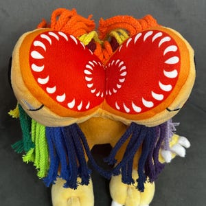 May include: A plush toy featuring a yellow body with two large, circular red mouth-like structures on the head, each lined with white teeth. The toy has multicolored yarn fringe in orange, yellow, green, blue, and purple, and white claws on its feet.