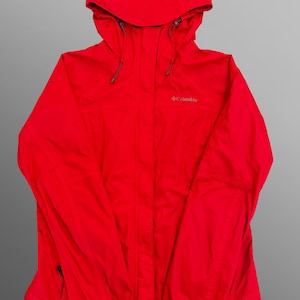 Puede incluir: Una chaqueta de lluvia roja brillante de Columbia con capucha y cordones ajustables, cierre frontal de cremallera oculto por una solapa y mangas largas con puños ajustables. El nombre de la marca Columbia está bordado en gris en el pecho.
