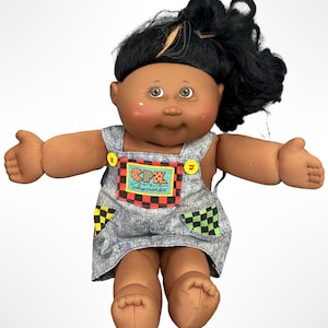 May include: A Cabbage Patch Kids doll with dark skin, brown eyes, and black hair styled in a messy ponytail. The doll wears denim-style overalls featuring a front pocket with a red and black checkered border, yellow buttons, and a CPK logo patch.