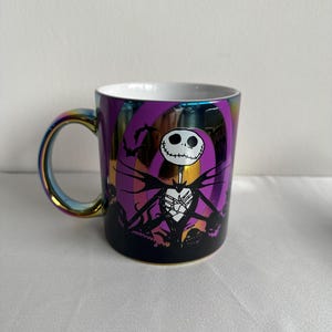 Pode incluir: Uma caneca de cer&acirc;mica com acabamento iridescente apresentando um gr&aacute;fico de um personagem esquel&eacute;tico em um terno risca de giz contra um fundo espiral roxo. O design inclui silhuetas pretas de morcegos e ab&oacute;boras, com interior e al&ccedil;a brancos.