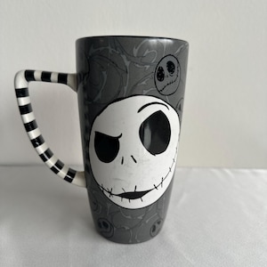 Pode incluir: Uma caneca alta de cer&acirc;mica com fundo cinza e um padr&atilde;o repetitivo de rostos de Jack Skellington. A parte frontal exibe um rosto de caveira branco grande e proeminente com olhos pretos e uma boca costurada. A al&ccedil;a tem um design listrado em preto e branco.