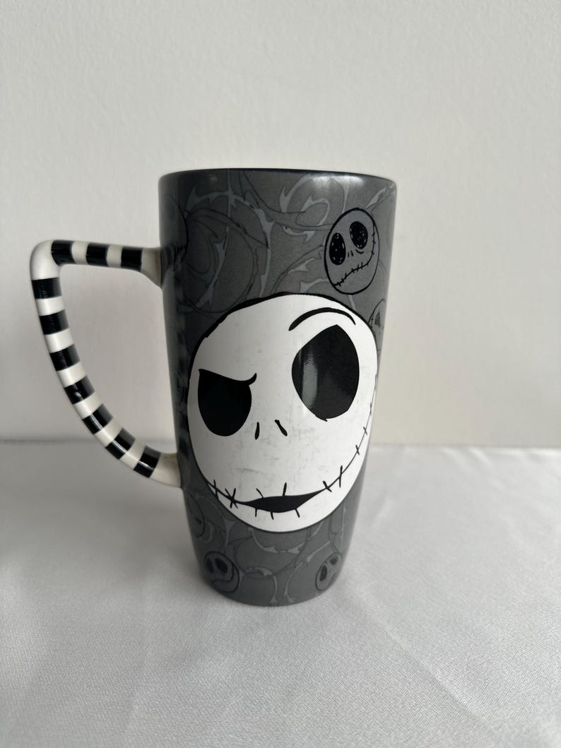 Pode incluir: Uma caneca alta de cer&acirc;mica com fundo cinza e um padr&atilde;o repetitivo de rostos de Jack Skellington. A parte frontal exibe um rosto de caveira branco grande e proeminente com olhos pretos e uma boca costurada. A al&ccedil;a tem um design listrado em preto e branco.