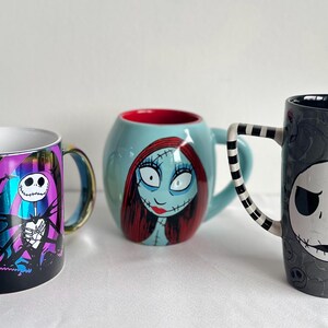 Pode incluir: Tr&ecirc;s canecas de cer&acirc;mica com personagens de O Estranho Mundo de Jack. A caneca da esquerda &eacute; roxa e preta com Jack Skellington. A caneca central &eacute; azul clara com Sally. A caneca da direita &eacute; alta, cinza e apresenta o rosto de Jack.