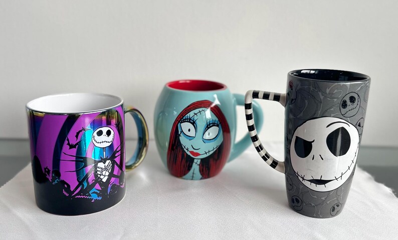 Pode incluir: Tr&ecirc;s canecas de cer&acirc;mica com personagens de O Estranho Mundo de Jack. A caneca da esquerda &eacute; roxa e preta com Jack Skellington. A caneca central &eacute; azul clara com Sally. A caneca da direita &eacute; alta, cinza e apresenta o rosto de Jack.