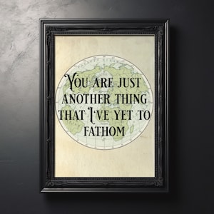 May include: A framed print with a vintage map design. The map is circular, with green landmasses and a cream-colored background. The text reads, "You are just another thing that I've yet to fathom." The frame is black.
