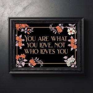 Könnte beinhalten: Schwarz gerahmter Kunstdruck mit dem Zitat "YOU ARE WHAT YOU LOVE, NOT WHO LOVES YOU" in goldfarbenen Buchstaben. Der Druck ist mit orangefarbenen, weißen und lila floralen Akzenten verziert.