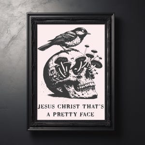 Könnte beinhalten: Schwarz-weiß gerahmter Kunstdruck mit einem Totenkopf, aus dem Pilze wachsen, und einem Vogel, der obenauf sitzt. Der Text "JESUS CHRIST THAT'S A PRETTY FACE" ist unten auf dem Bild aufgedruckt.