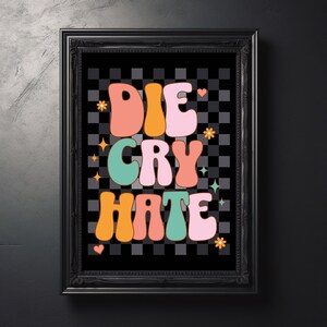 May include: A framed art print with the words "DIE CRY HATE" in colorful, retro-style lettering. The background is a black and gray checkered pattern, with decorative stars, hearts, and flowers. The frame is black.