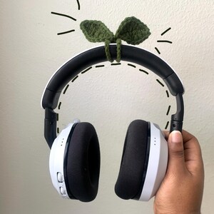 May include: White and black wireless headphones featuring a dark green crocheted leaf attachment on the headband. The accessory is secured with a matching green yarn loop, and the image includes hand-drawn dashed lines around the headset.