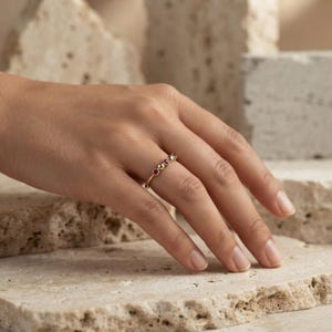 May include: A thin gold ring featuring a delicate arrangement of small, round, multi-colored gemstones and a single polished gold bead set along the band. The ring is worn on a finger, showcasing the fine metalwork and the vibrant, varied colors of the stones.
