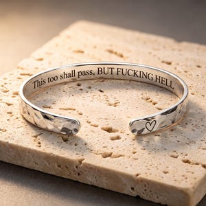 May include: A silver-toned metal cuff bracelet with a hammered, textured exterior. The interior is engraved with the black text "This too shall pass, BUT FUCKING HELL" and a small heart symbol is etched near one of the open ends of the bracelet.