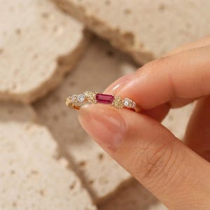 May include: A gold ring featuring a central baguette-cut red gemstone flanked by two small clear round stones on each side. The band is accented with delicate metal leaf motifs surrounding the stones, creating a detailed and ornate appearance.