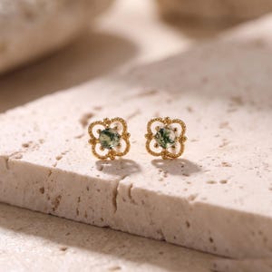May include: A pair of gold stud earrings featuring a round moss agate gemstone set within a decorative, openwork four-leaf clover frame. The metal has a polished finish, and the stones display natural green inclusions within the translucent surface.