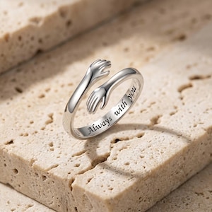 Puede incluir: Un anillo abierto de tono plateado que presenta dos manos envolviendo la banda en un gesto de abrazo. La superficie interior de la banda está grabada con el texto negro Always with you en una tipografía cursiva.