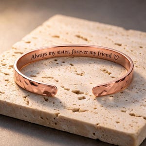 May include: A rose gold-toned metal cuff bracelet with a hammered, textured exterior. The inner surface is engraved with the black text "Always my sister, forever my friend" followed by a small heart symbol. The bracelet has an open design.