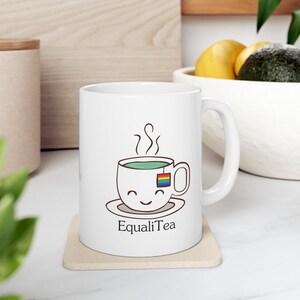 Può includere: Tazza in ceramica bianca con un'illustrazione di cartone animato di una tazza di tè con una bustina di tè arcobaleno. La tazza di tè ha un viso sorridente e il testo "EqualiTea" è stampato sotto la tazza di tè.