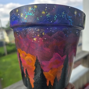 Op de afbeelding: Een cilindervormige plantenpot met een handgeschilderde nachthemel. Het ontwerp verloopt van een donkerblauwe sterrenhemel met glitteraccenten naar een levendige magenta en oranje zonsondergang, afgewerkt met zwarte silhouetten van dennenbomen.