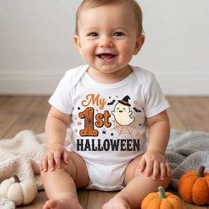 Puede incluir: Un body blanco de manga corta para bebé con un estampado gráfico que incluye el texto My 1st HALLOWEEN en naranja y negro. El diseño presenta un número uno con patrón de telaraña, un pequeño fantasma con sombrero negro y murciélagos.