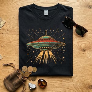 Puede incluir: Una camiseta negra de cuello redondo con un estampado gráfico de un platillo volante de estilo retro que emite un haz de luz amarillo, rodeado de pequeñas estrellas. El diseño utiliza tonos rojos, azul verdoso y amarillos sobre la tela.