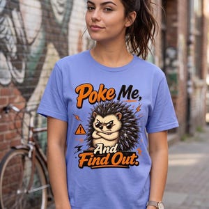 Puede incluir: Una camiseta de color morado claro con el gráfico de un erizo gruñón con gafas de sol y los brazos cruzados. El texto "Poke Me, And Find Out" aparece en letras naranjas, junto a un icono de triángulo de advertencia amarillo y rayos.