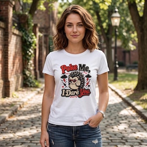Puede incluir: Una camiseta blanca de manga corta con un gráfico de un erizo con gafas de sol en forma de corazón y el texto "Poke Me, I Dare You". El diseño incluye rayos rojos, nubes negras y estrellas que rodean al personaje del erizo.