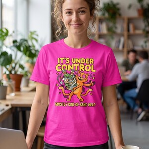 Puede incluir: Una camiseta de manga corta de color rosa brillante con un gato naranja de dibujos animados que sostiene una pila de papeles y tazas de café. El texto IT'S UNDER CONTROL aparece arriba y MOSTLY KIND OF SEND HELP abajo, con estrellas amarillas.